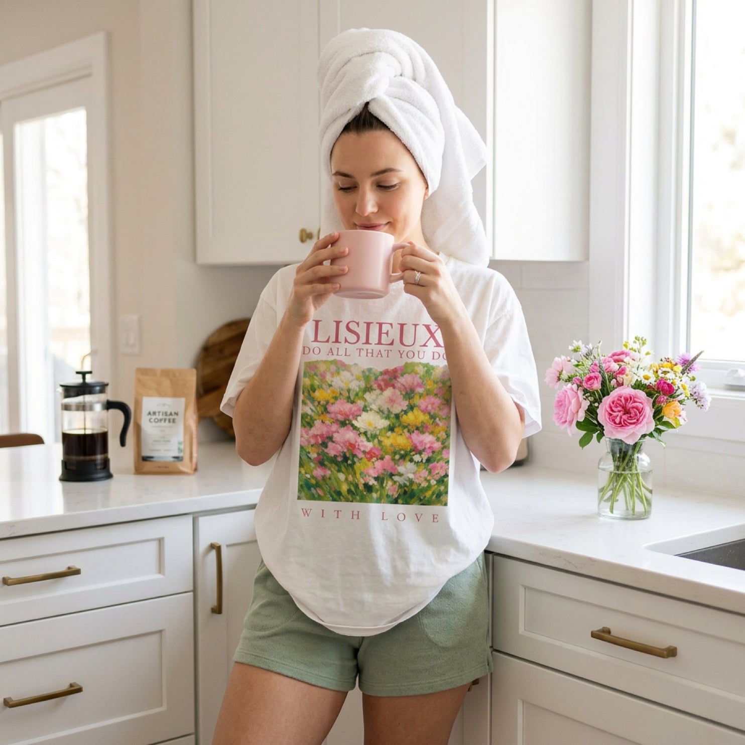 Woman in towel and "Lisieux with love" shirt holding pink mug by flowers.