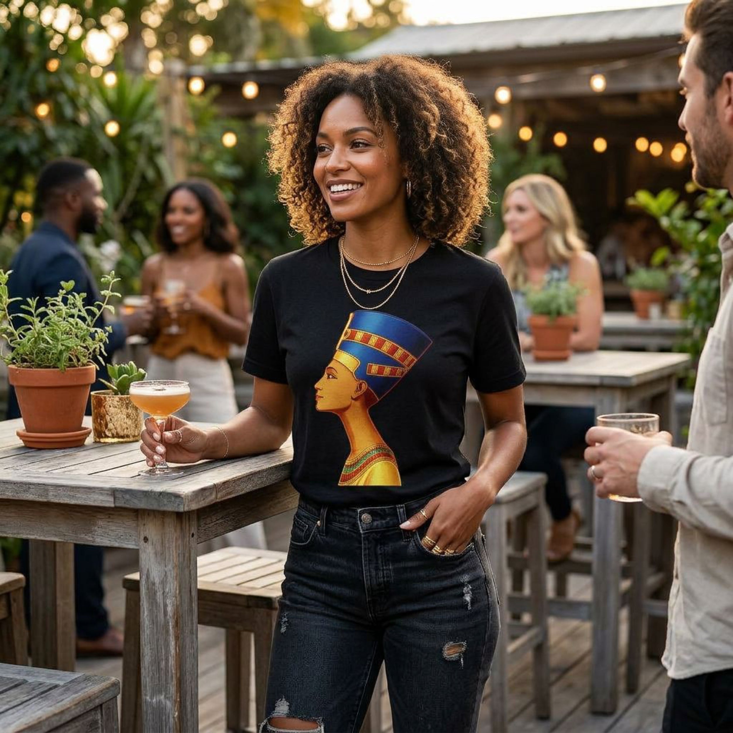 Woman in black t-shirt with Nefertiti graphic, holding cocktail outdoors.