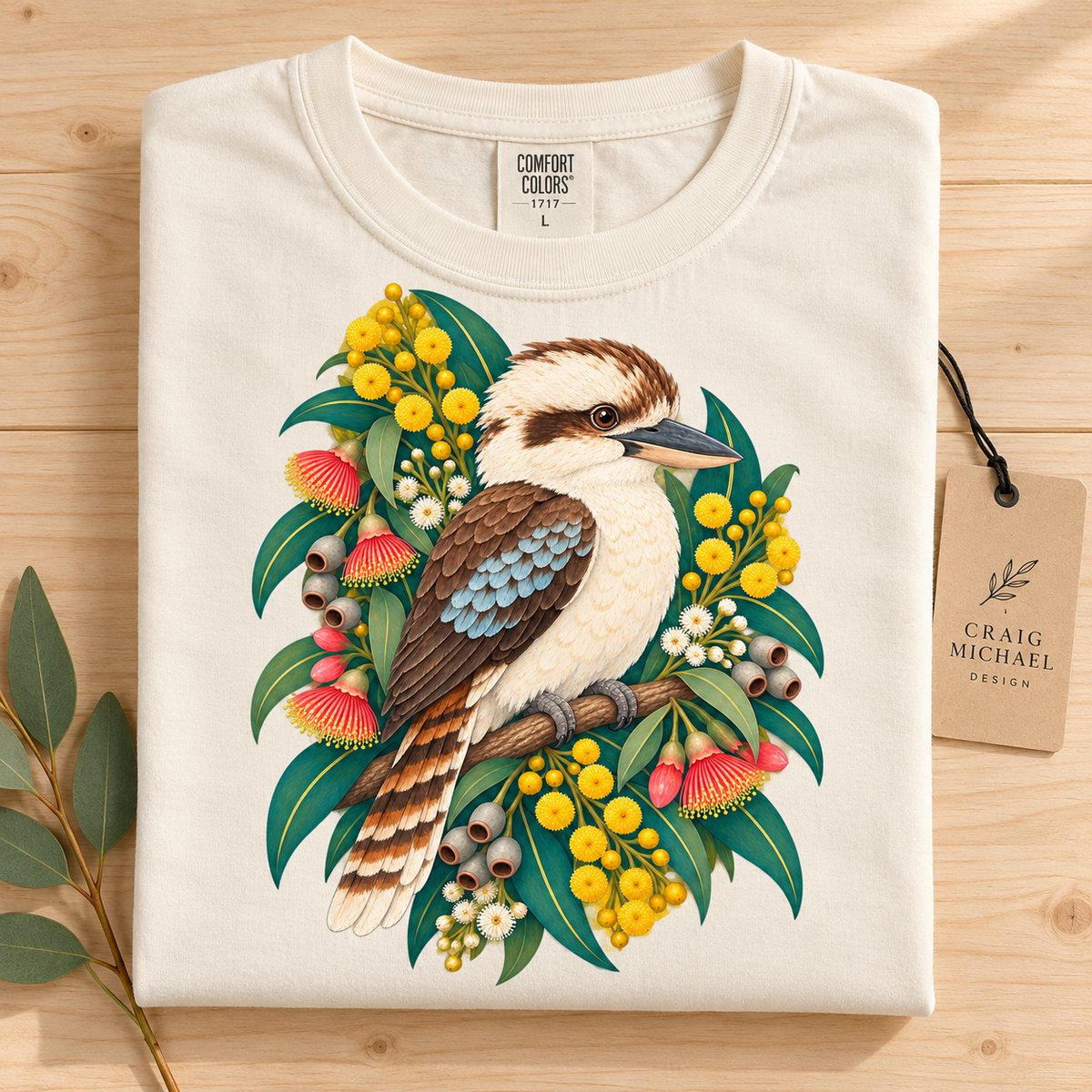 Cream t-shirt folded on wood, featuring a kookaburra among Australian wildflowers and gum leaves.