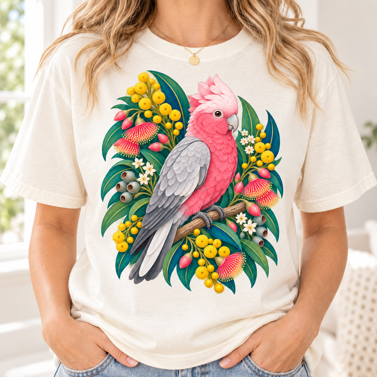 A person wearing a cream-colored t-shirt printed with a pink and grey cockatoo surrounded by colorful tropical foliage.