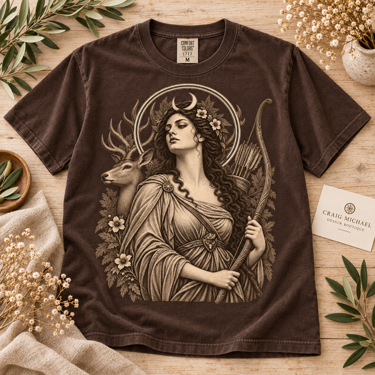 Brown t-shirt with a detailed illustration of a woman with a crescent moon crown, bow, and arrows, next to a deer.