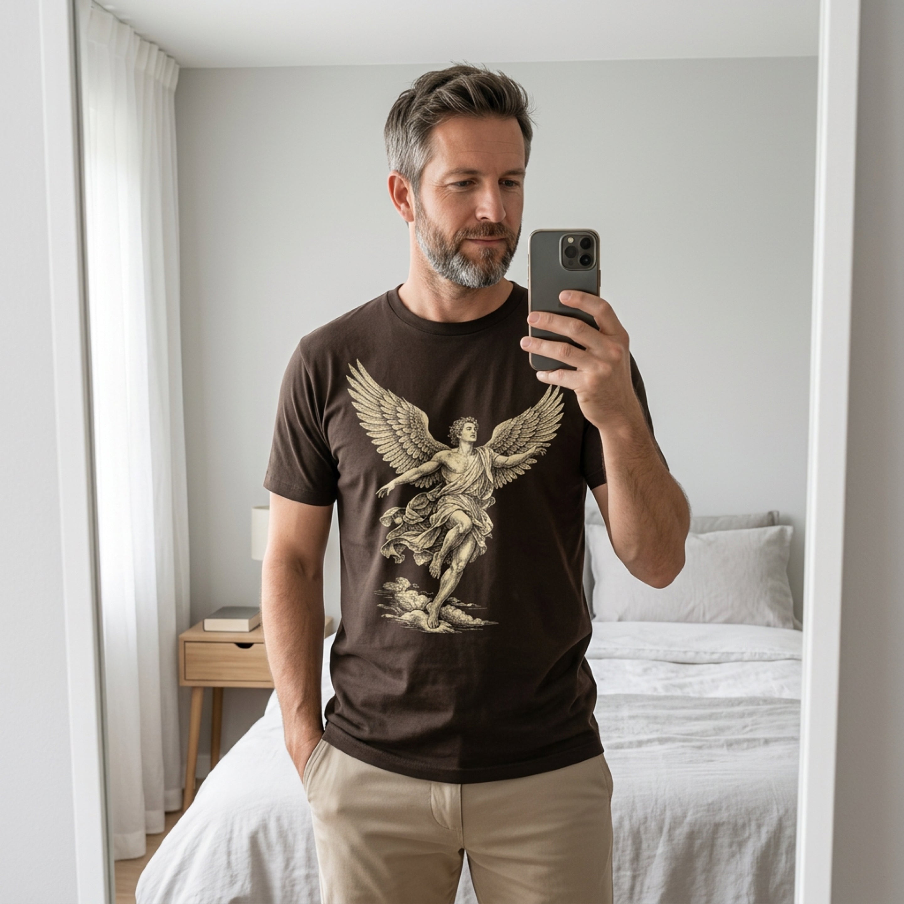 Man in brown t-shirt with angelic graphic, taking mirror selfie in bedroom.