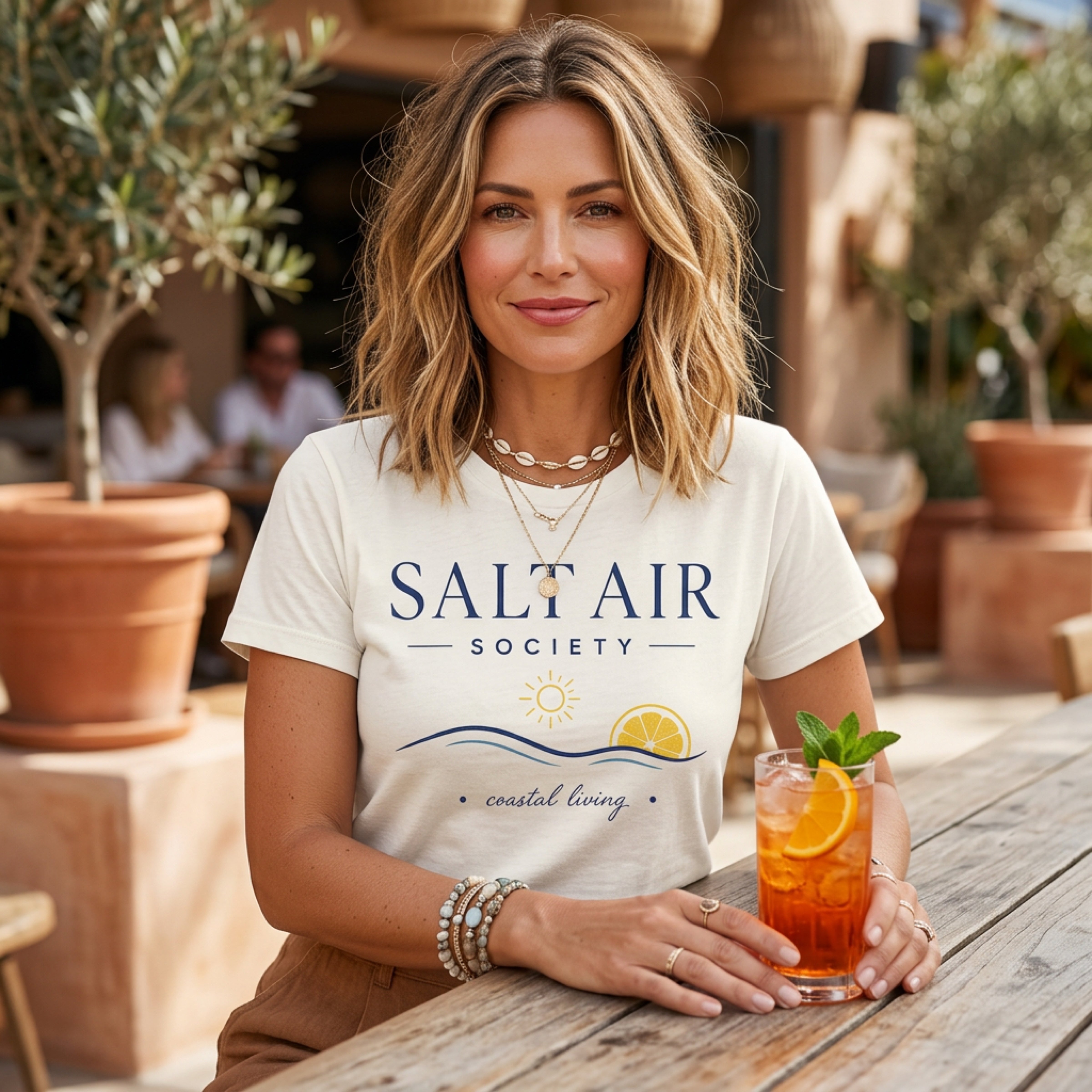 Woman in "Salt Air Society" t-shirt holding an orange drink outdoors.