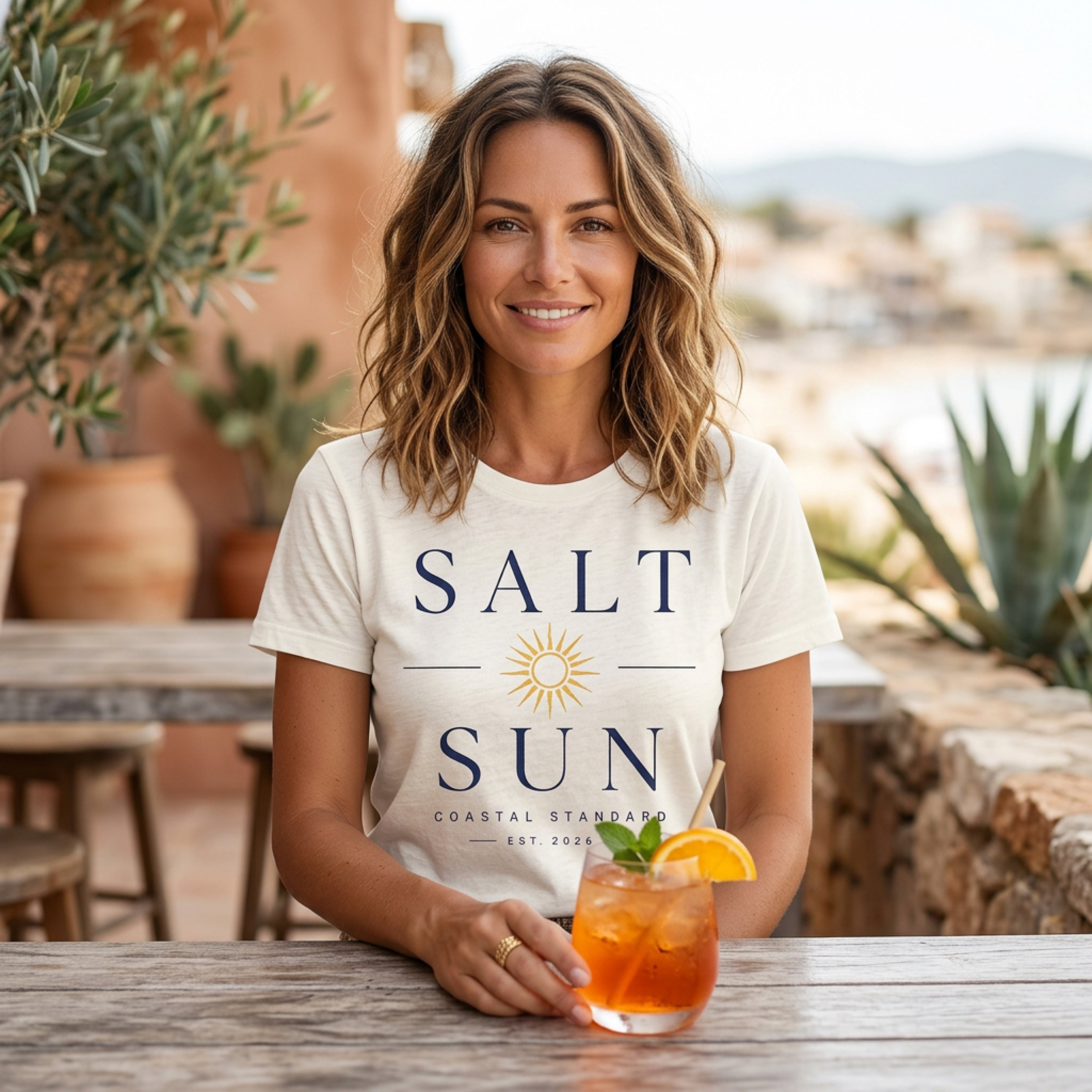 Woman wearing a cream t-shirt with "SALT SUN" logo, holding a cocktail.