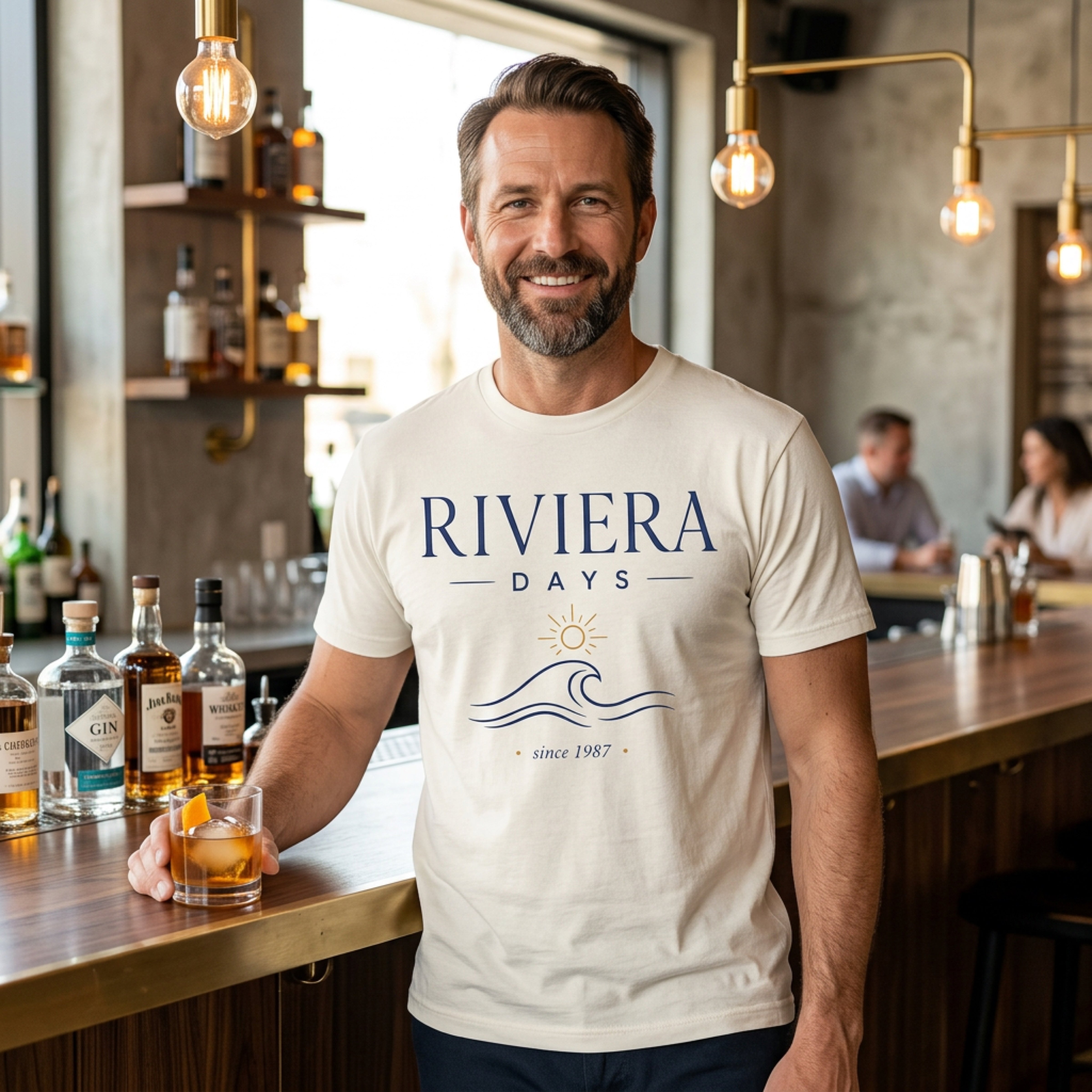 Man in cream t-shirt with "Riviera Days Since 1987" text, holding a drink.
