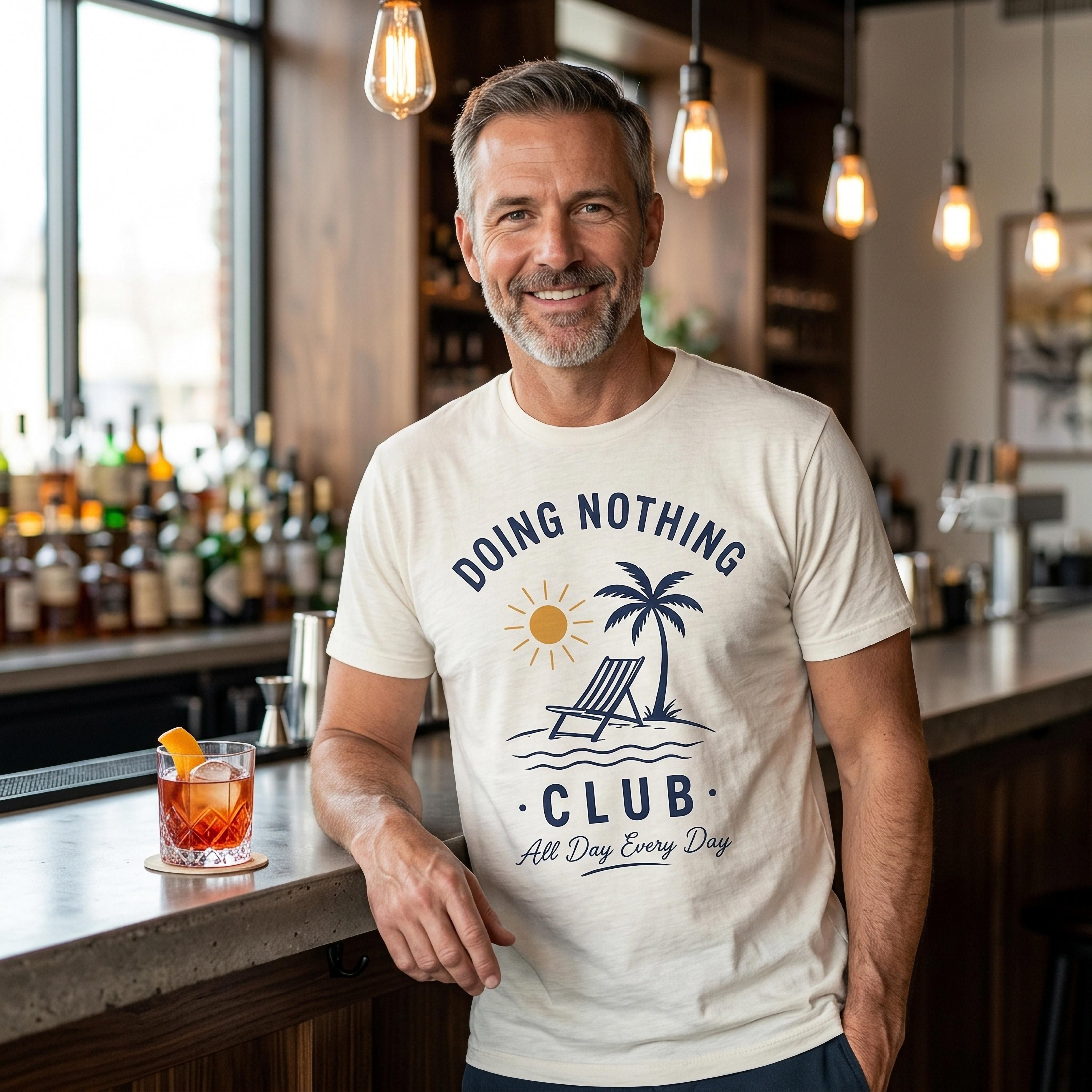 Man in cream t-shirt with "Doing Nothing Club" sunset graphic leans on a bar.