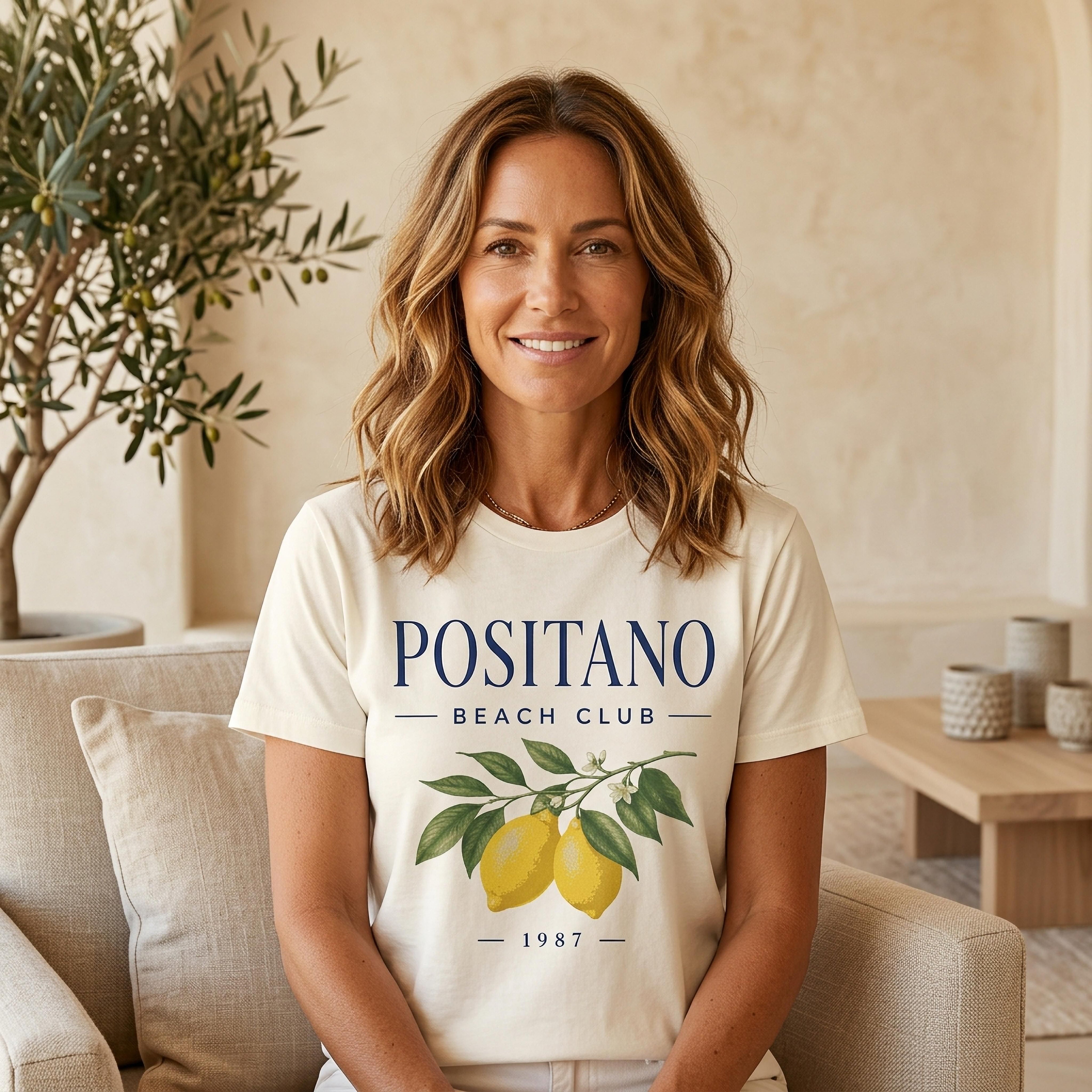 Woman wearing a cream t-shirt with "Positano Beach Club 1987" and lemon illustration.
