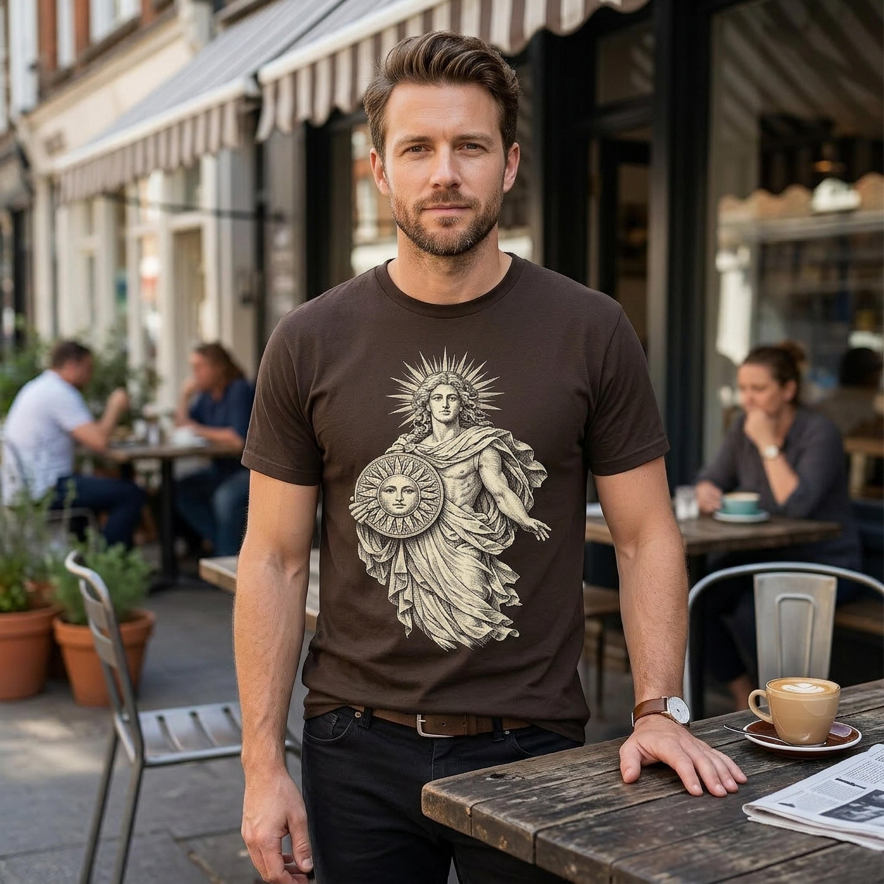 Man wearing brown t-shirt with sun and goddess graphic, standing at outdoor cafe table.