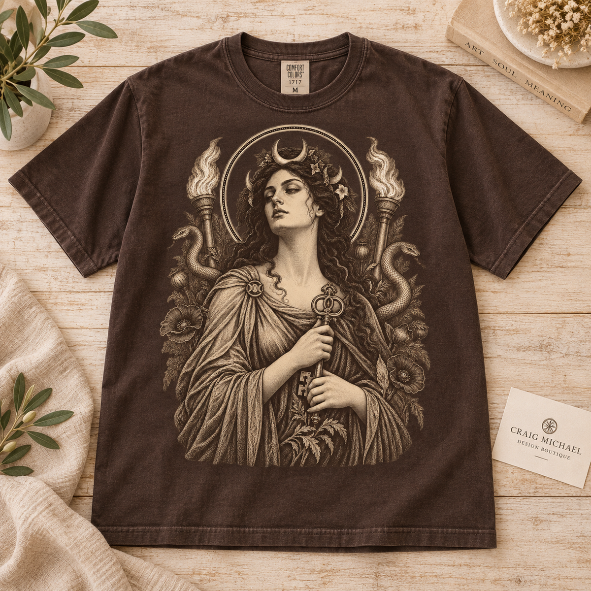 Brown t-shirt featuring a classical illustration of a woman holding a key, with torches and snakes.