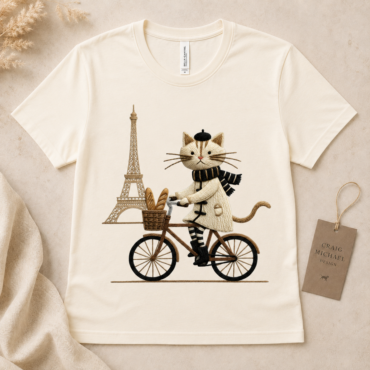 Cream t-shirt with embroidered cat riding bicycle in front of Eiffel Tower.